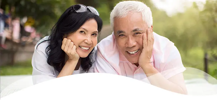 Smiling older couple lies on grass with heads propped on hands, exuding warmth and happiness. Lush greenery and soft light create a serene atmosphere.